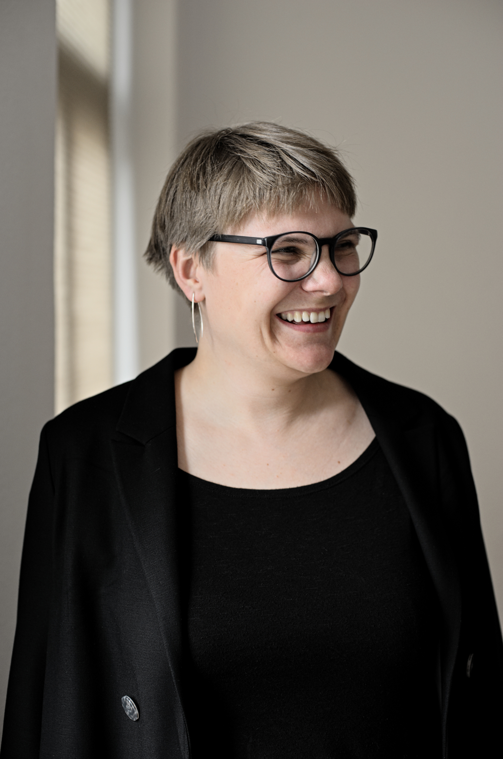 Ein Portrait von Luise Veit. Es ist ihr Oberkörper zu sehen.  Sie schaut zur Seite und lacht. Luise Veit hat kurze Haare und eine Brille mit schwarzem Rand. Sie trägt silberne Ohrringe, ein schwarzes Oberteil und einen schwarzen Blazer.